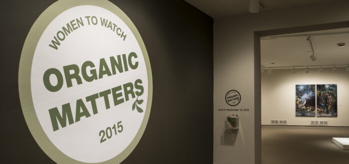 View of a gallery space. In big letters green , the text on the wall reads "Women to Watch: Organic Matters, 2015."