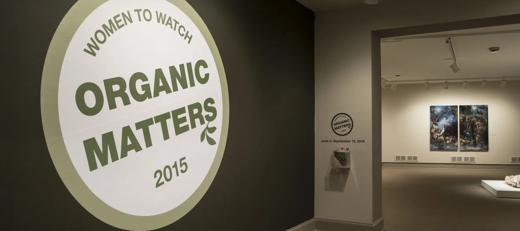 View of a gallery space. In big letters green , the text on the wall reads "Women to Watch: Organic Matters, 2015."