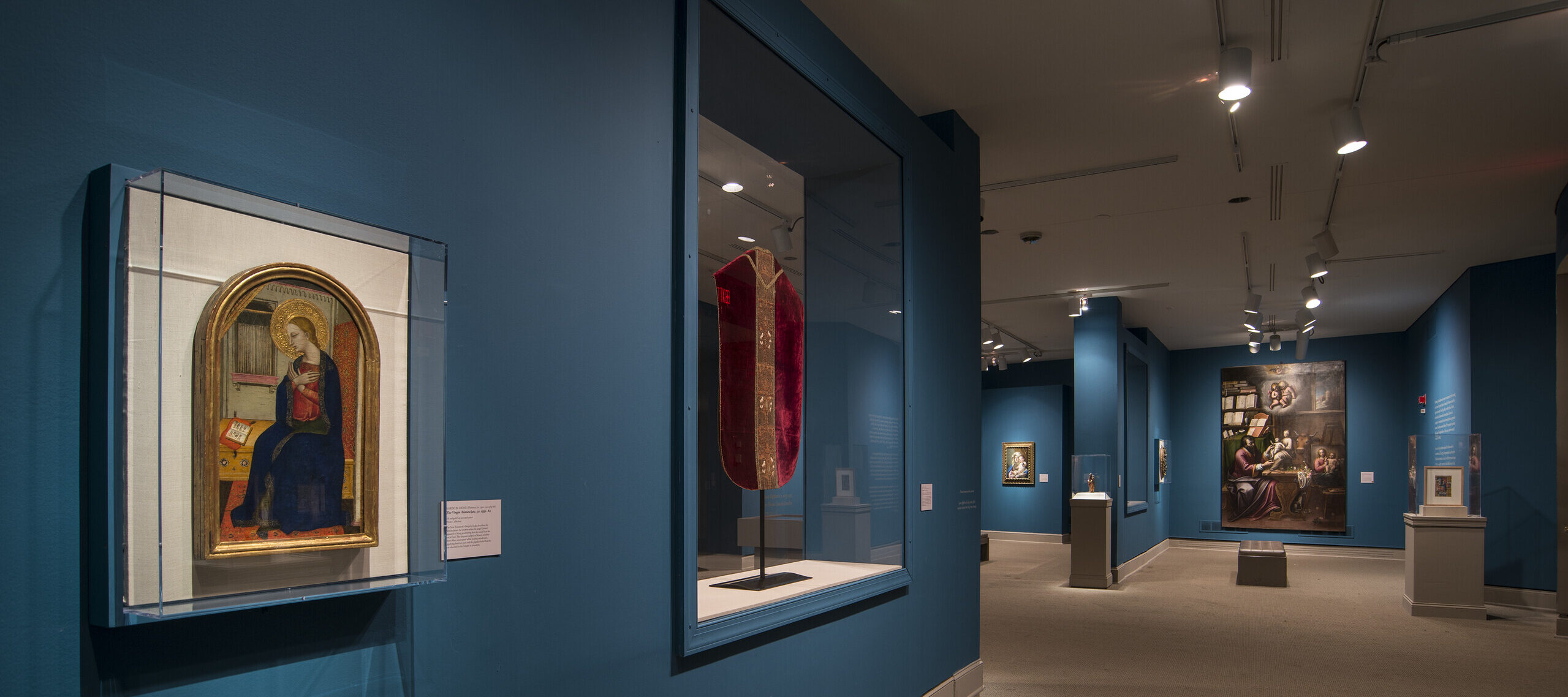 View of a gallery space with blue walls. On the left is a painting of a woman in a dark blue dress in a golden frame. Next to it is a red cloth framed in glass.