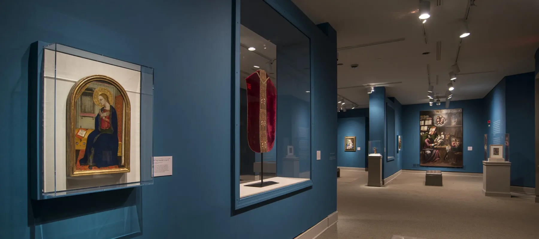 View of a gallery space with blue walls. On the left is a painting of a woman in a dark blue dress in a golden frame. Next to it is a red cloth framed in glass.