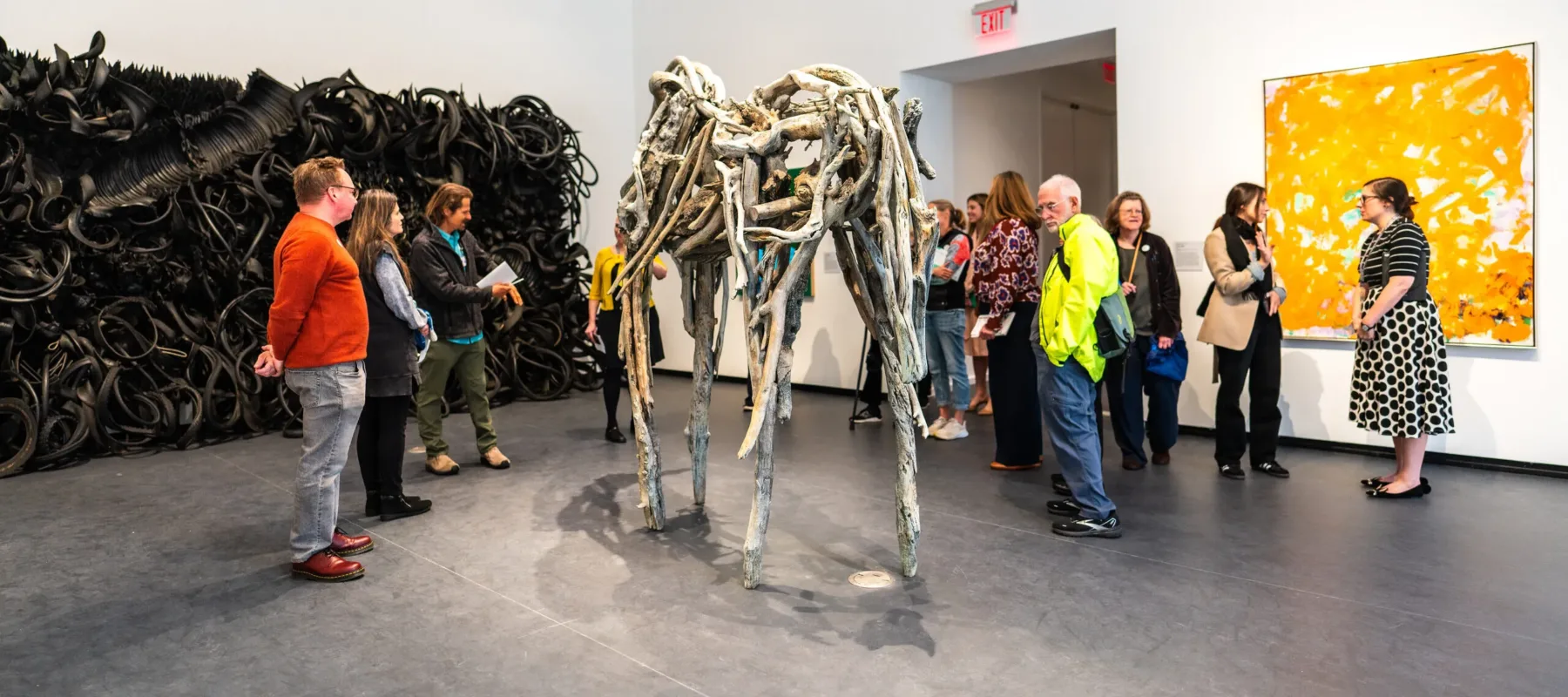 View of a gallery. Several people are standing in the space, viewing the artworks. A large, yellow painting hangs on the right wall, an even bigger sculpture made of rubber tires is on the left wall, and a sculpture made of branches stands in the middle of the room.