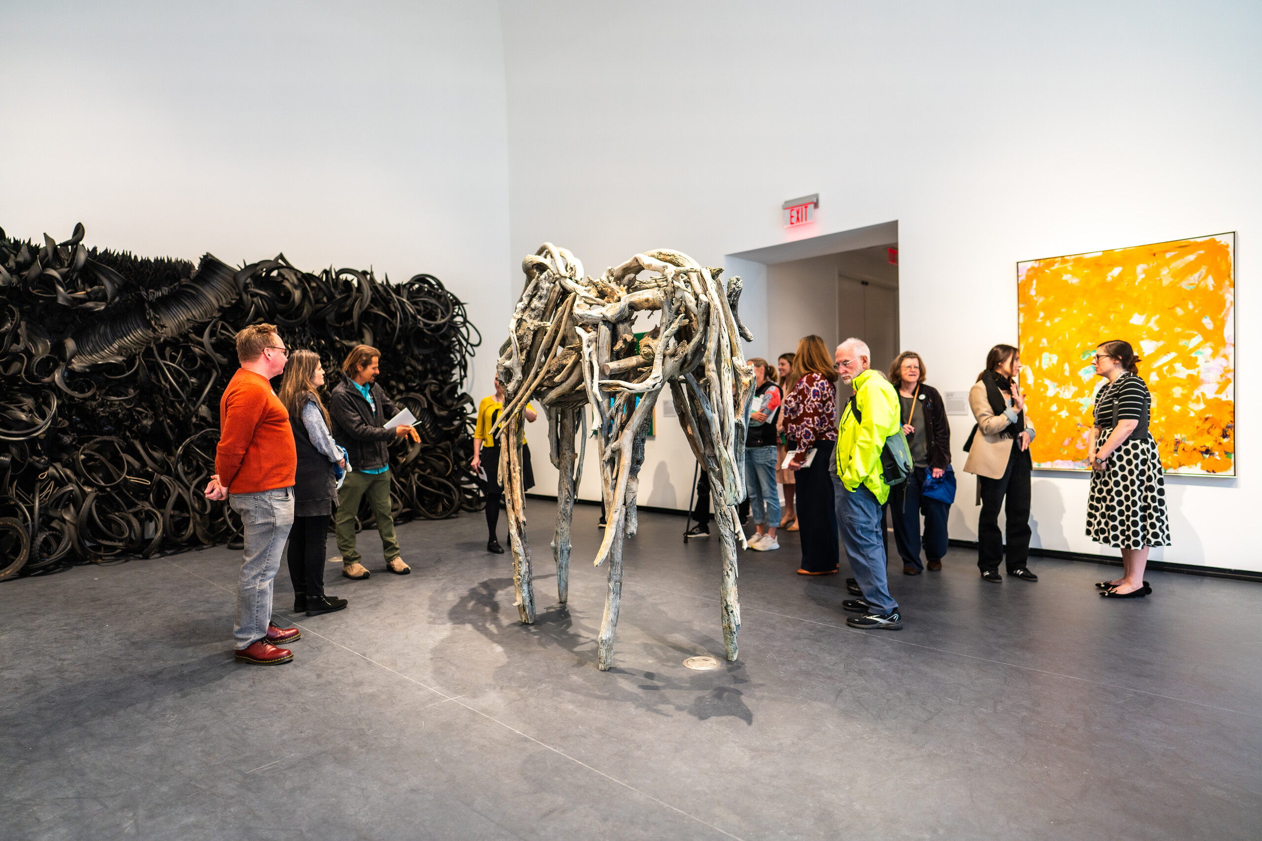 View of a gallery. Several people are standing in the space, viewing the artworks. A large, yellow painting hangs on the right wall, an even bigger sculpture made of rubber tires is on the left wall, and a sculpture made of branches stands in the middle of the room.