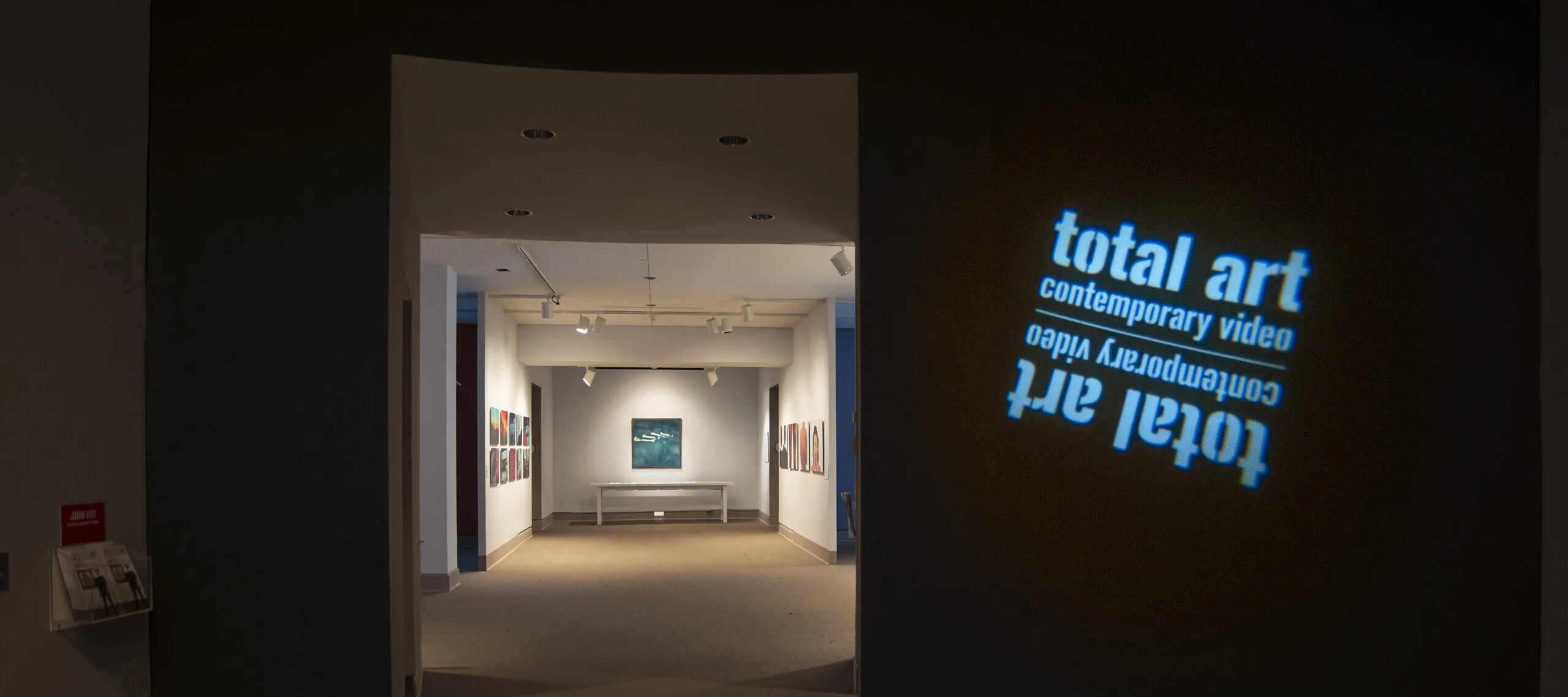 View of a gallery space. In a dark room, the title of the exhibition, "Total Art: Contemporary Video", is projected on a wall.