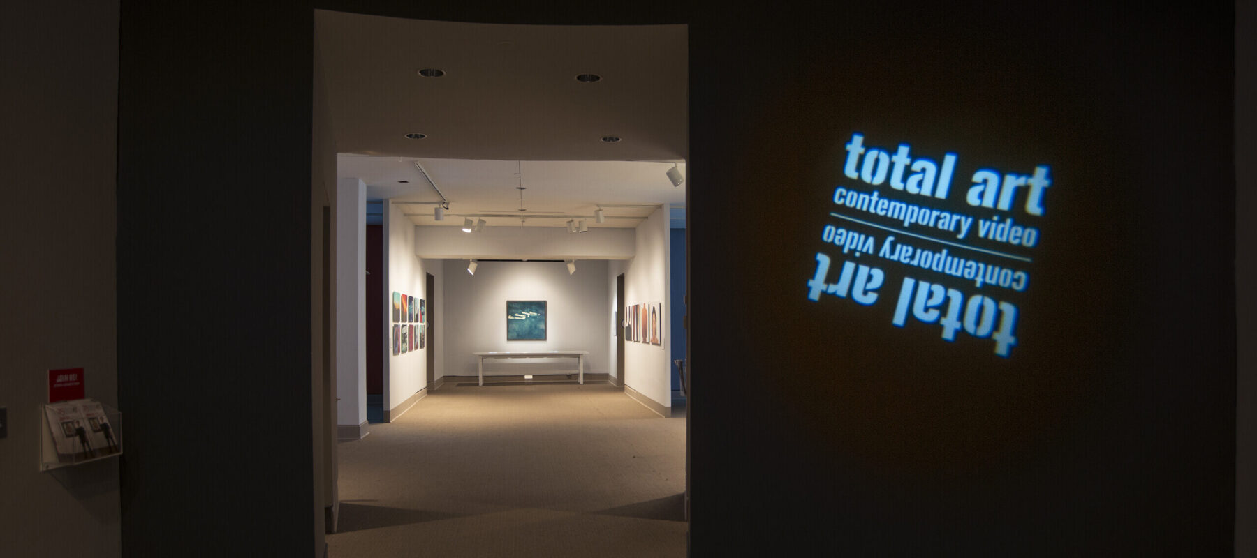 View of a gallery space. In a dark room, the title of the exhibition, "Total Art: Contemporary Video", is projected on a wall.