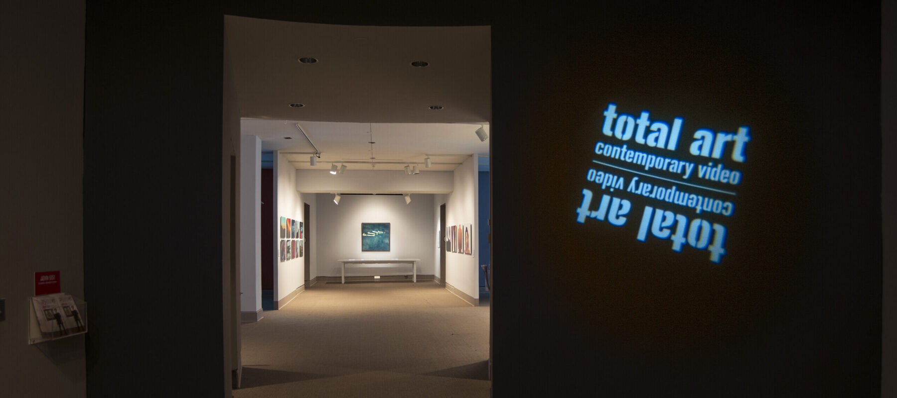 View of a gallery space. In a dark room, the title of the exhibition, "Total Art: Contemporary Video", is projected on a wall.