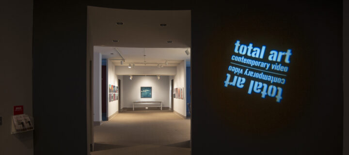 View of a gallery space. In a dark room, the title of the exhibition, "Total Art: Contemporary Video", is projected on a wall.