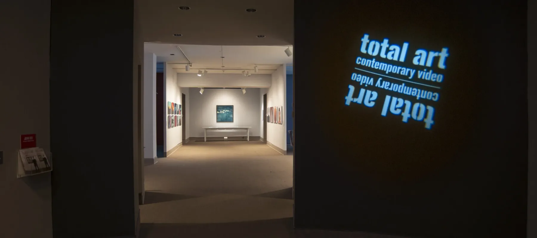 View of a gallery space. In a dark room, the title of the exhibition, "Total Art: Contemporary Video", is projected on a wall.