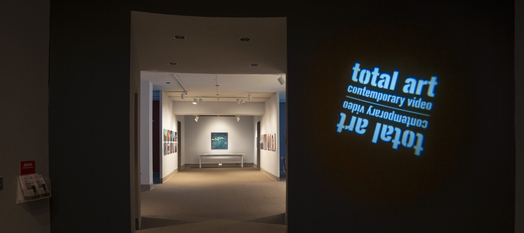 View of a gallery space. In a dark room, the title of the exhibition, "Total Art: Contemporary Video", is projected on a wall.