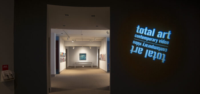 View of a gallery space. In a dark room, the title of the exhibition, "Total Art: Contemporary Video", is projected on a wall.