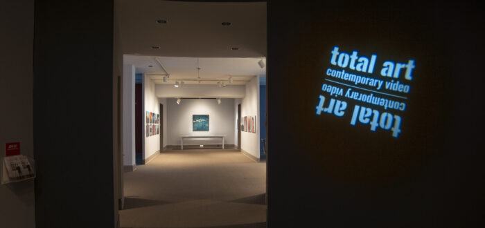View of a gallery space. In a dark room, the title of the exhibition, "Total Art: Contemporary Video", is projected on a wall.