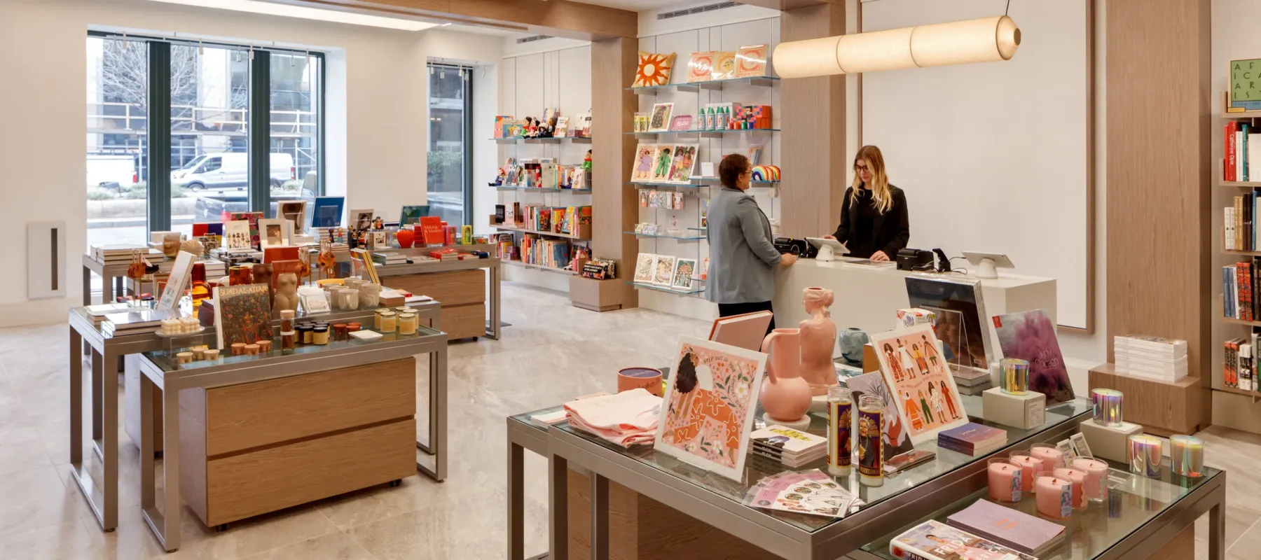 A museum gift shop is photographed at a wide angle. A colorful assortment of books, candles, textiles, and other small objects cover tables and shelves on the wall. A shopper is paying for items at a checkout table.