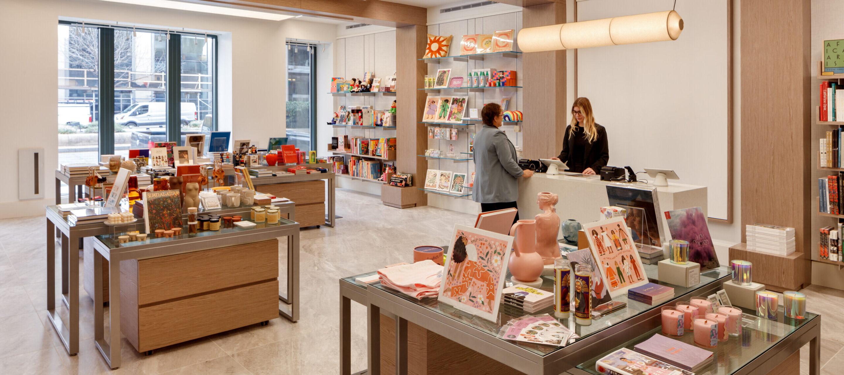 A museum gift shop is photographed at a wide angle. A colorful assortment of books, candles, textiles, and other small objects cover tables and shelves on the wall. A shopper is paying for items at a checkout table.