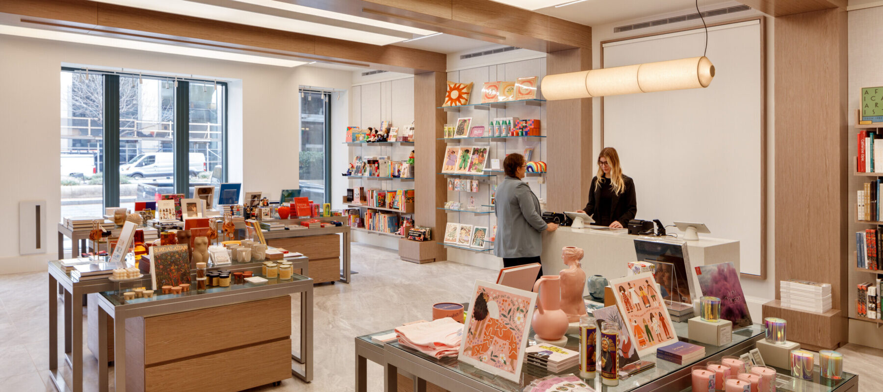 A museum gift shop is photographed at a wide angle. A colorful assortment of books, candles, textiles, and other small objects cover tables and shelves on the wall. A shopper is paying for items at a checkout table.