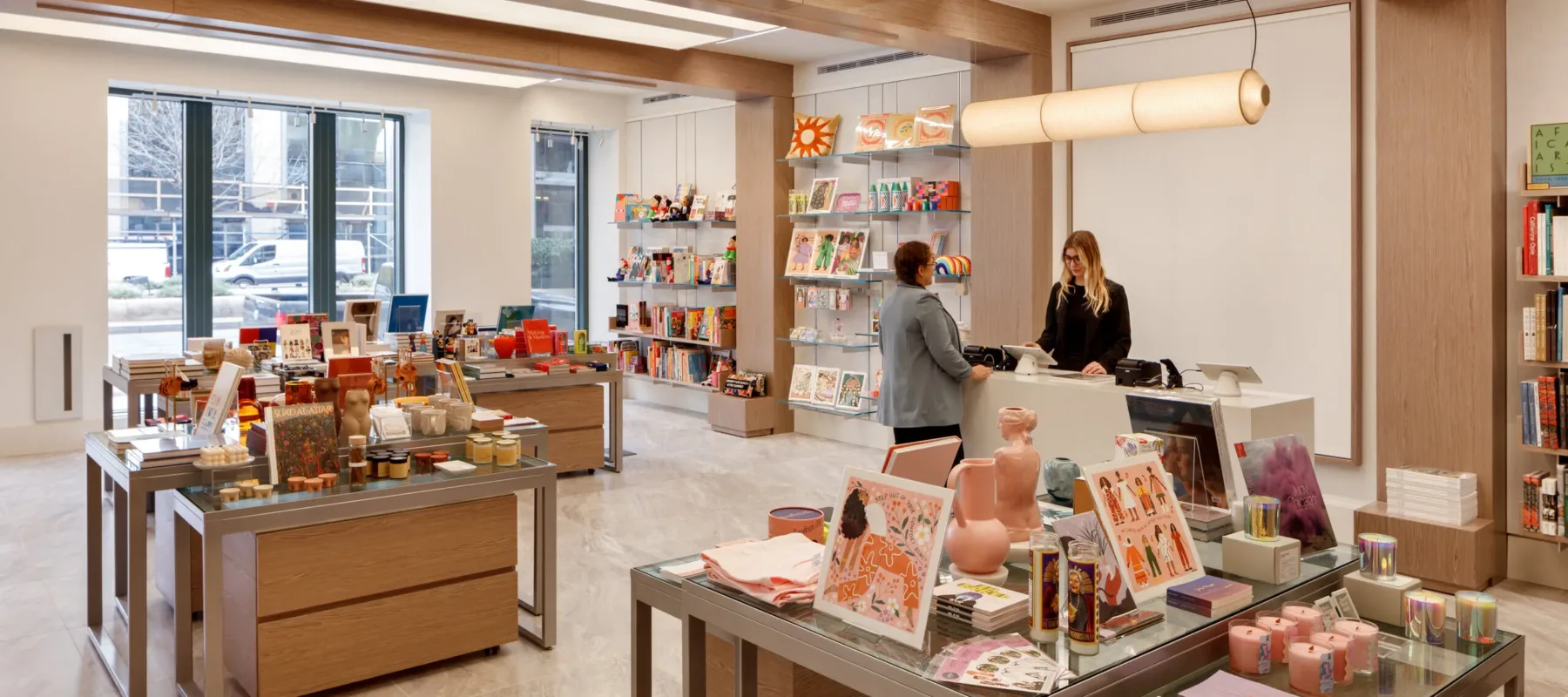 A museum gift shop is photographed at a wide angle. A colorful assortment of books, candles, textiles, and other small objects cover tables and shelves on the wall. A shopper is paying for items at a checkout table.