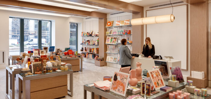 A museum gift shop is photographed at a wide angle. A colorful assortment of books, candles, textiles, and other small objects cover tables and shelves on the wall. A shopper is paying for items at a checkout table.