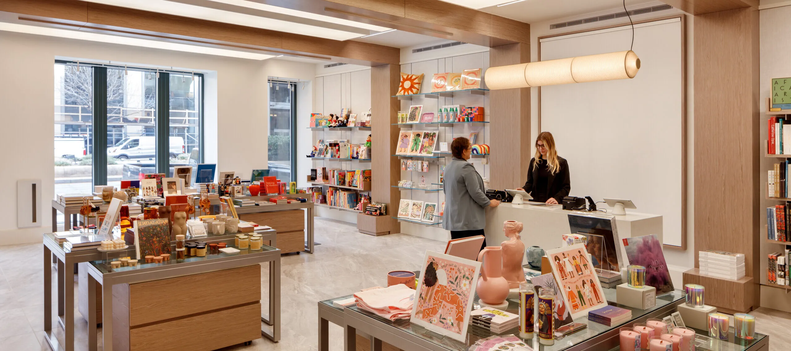 A museum gift shop is photographed at a wide angle. A colorful assortment of books, candles, textiles, and other small objects cover tables and shelves on the wall. A shopper is paying for items at a checkout table.