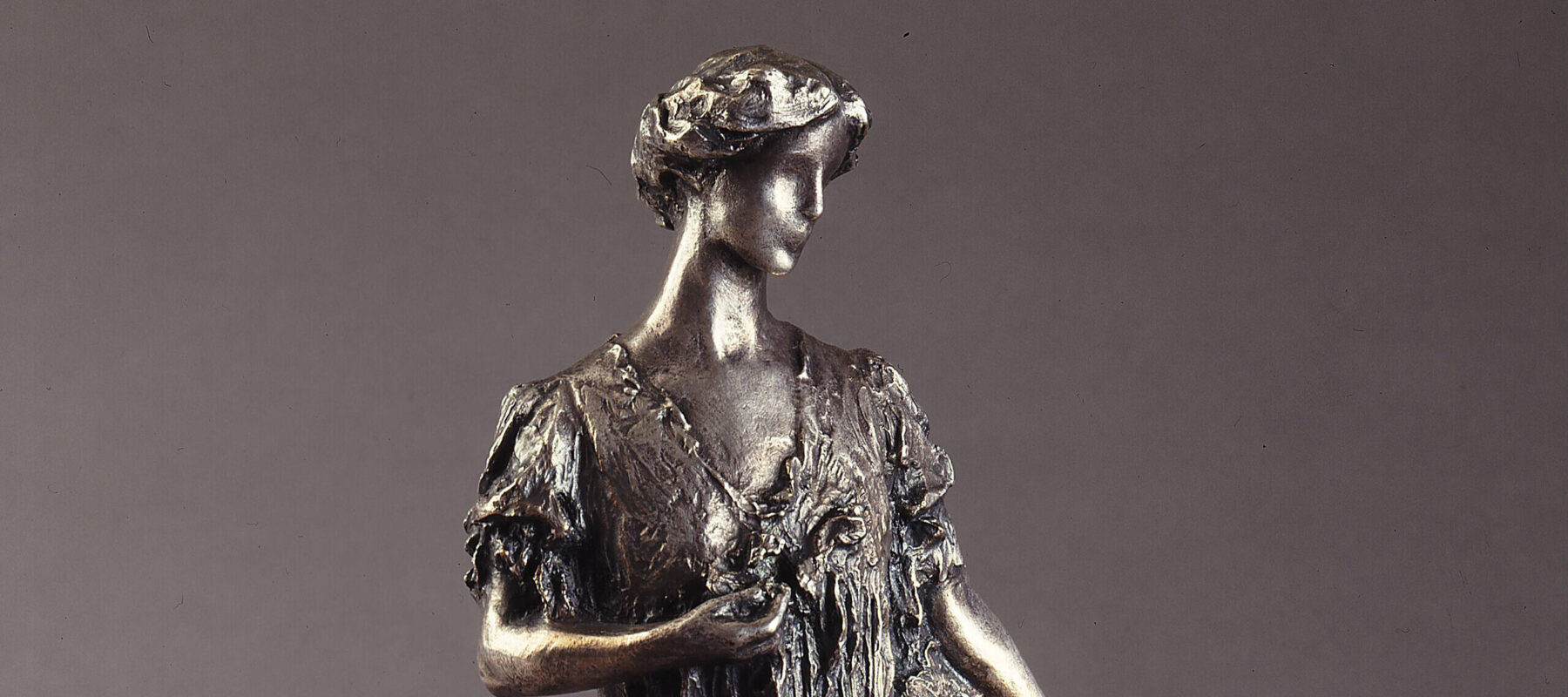 Bronze sculpture of a standing woman gazing over her right shoulder, holding a fan by her side. She wears a long, loose gown that gathers at her feet, with her hair swept off her neck. Her left hand holds a flower by her bust.
