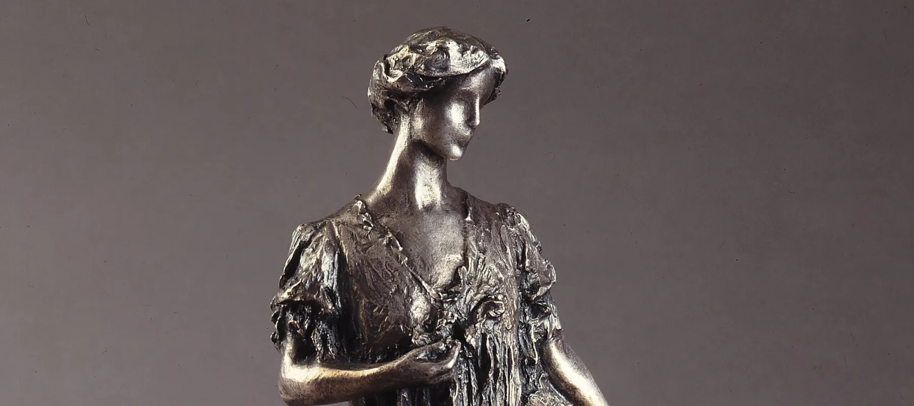 Bronze sculpture of a standing woman gazing over her right shoulder, holding a fan by her side. She wears a long, loose gown that gathers at her feet, with her hair swept off her neck. Her left hand holds a flower by her bust.