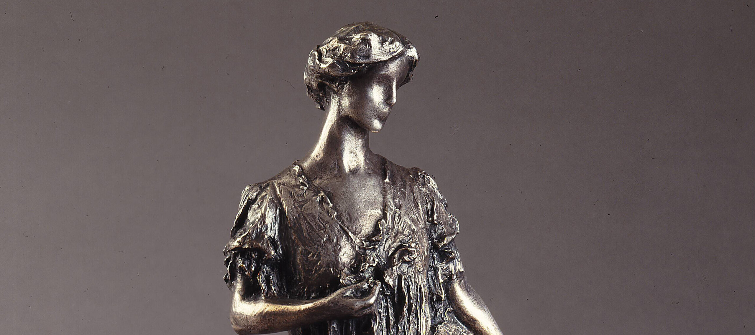 Bronze sculpture of a standing woman gazing over her right shoulder, holding a fan by her side. She wears a long, loose gown that gathers at her feet, with her hair swept off her neck. Her left hand holds a flower by her bust.