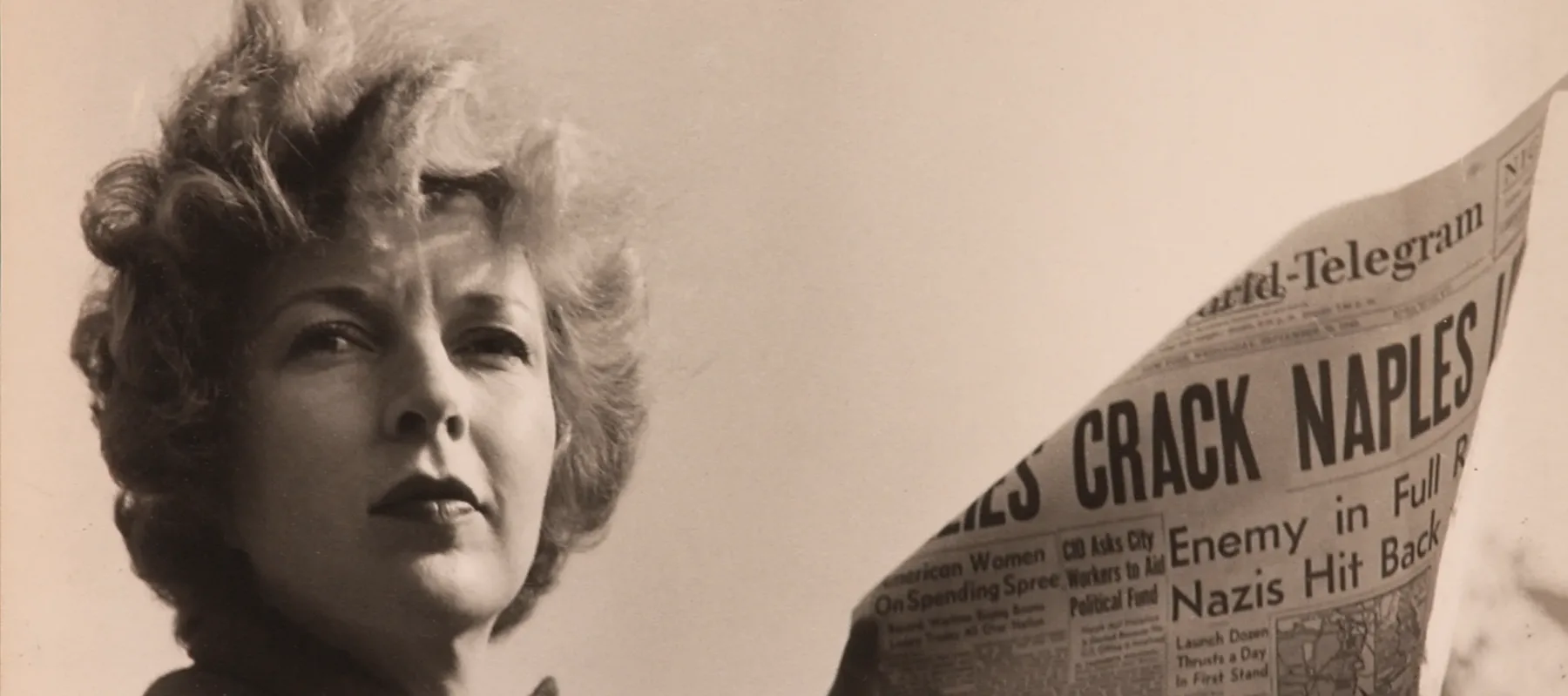 A black-and-white photograph of a light-skinned adult woman holding a newspaper with news about World War II. She wears a coat and her short, curly hair is caught in the wind.