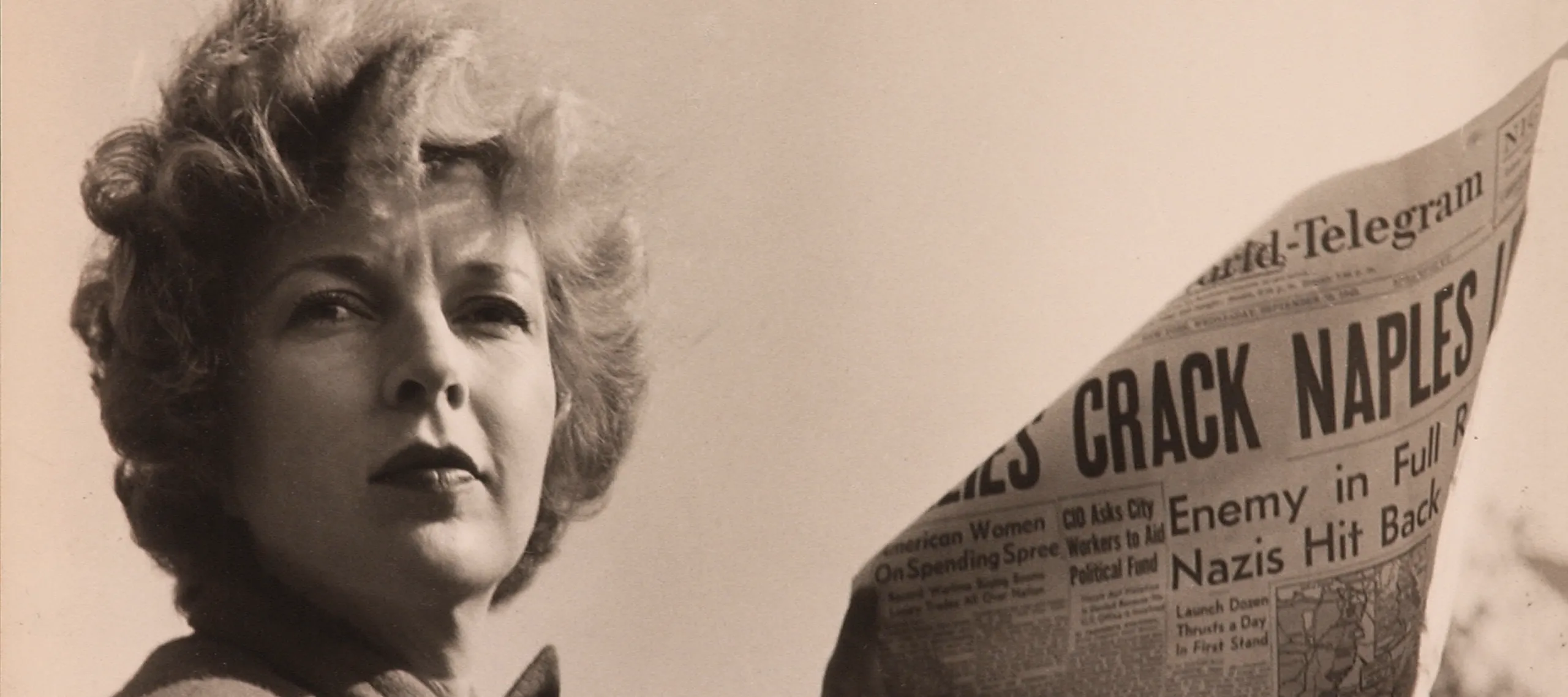 A black-and-white photograph of a light-skinned adult woman holding a newspaper with news about World War II. She wears a coat and her short, curly hair is caught in the wind.