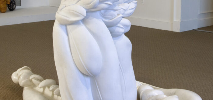 A photo of an abstract, white marble statue installed in a museum gallery. The sculpture appears to be a cluster of feathers with two large feathers resting on the ground.