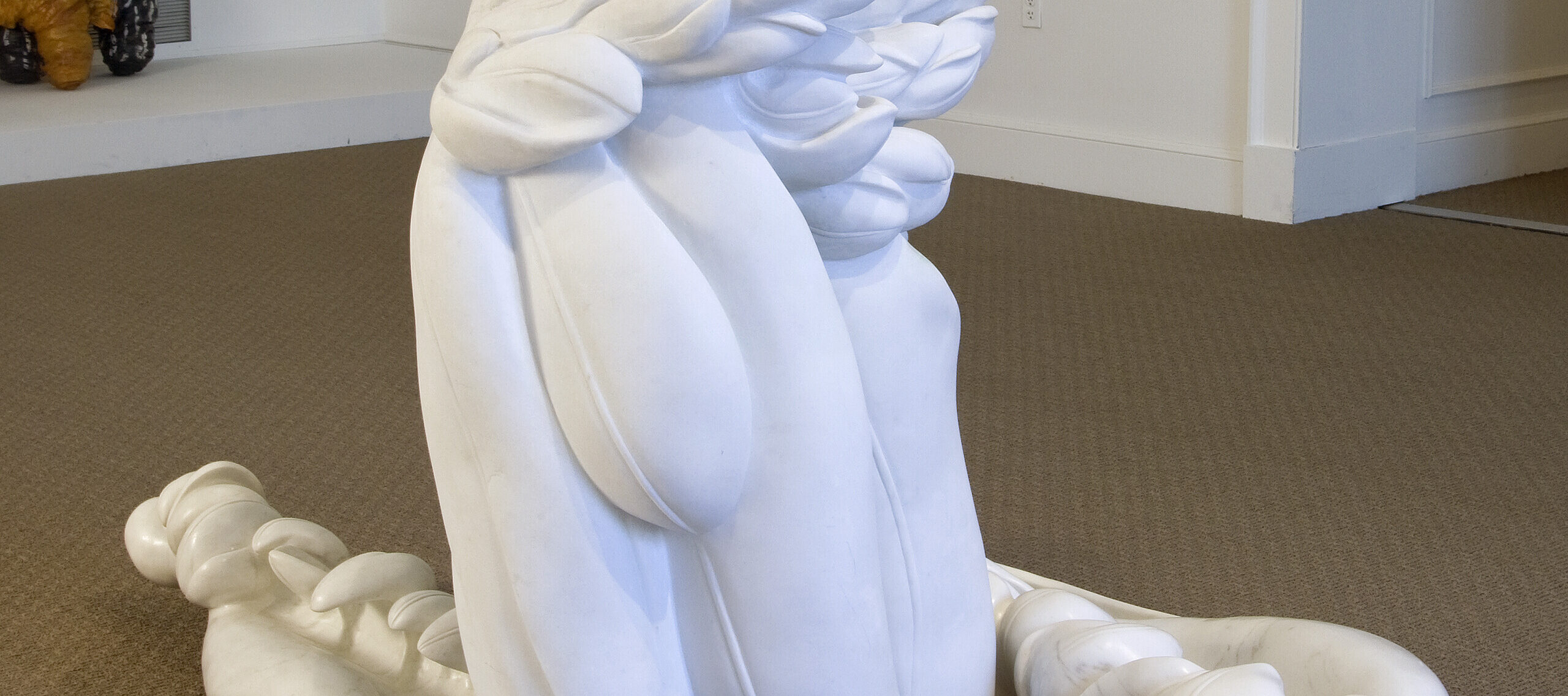 A photo of an abstract, white marble statue installed in a museum gallery. The sculpture appears to be a cluster of feathers with two large feathers resting on the ground.