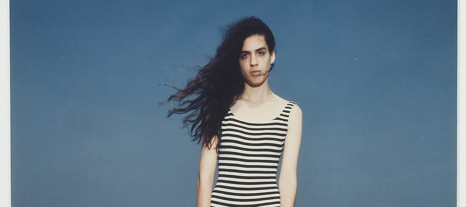 A light-skinned person wearing a black and white striped bathing suit standing at the beach with their back turned towards the ocean. Their long dark hair is blowing in the wind and their arms are held straight at their sides.