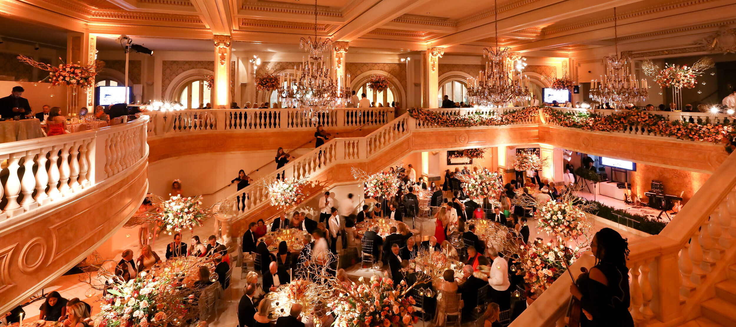 View of the gala from above looking out at the chandeliers, violin players on the marble staircase, and people gathered at round tables surrounded by abundant floral arrangements.