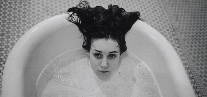 A black-and-white photograph of a light-skinned girl submerged in a white bathtub. Only her head is visible above the soap suds, and her dark hair hangs over the side of the tub. The floor beneath the tub is tiled.