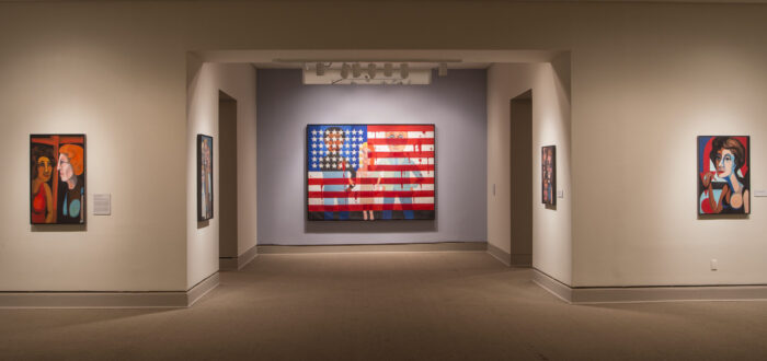 Installation view of a gallery space. A large painting showing an American flag splattered in blood and with people with a light skin tone standing behind the flag is hanging in the middle of the room.
