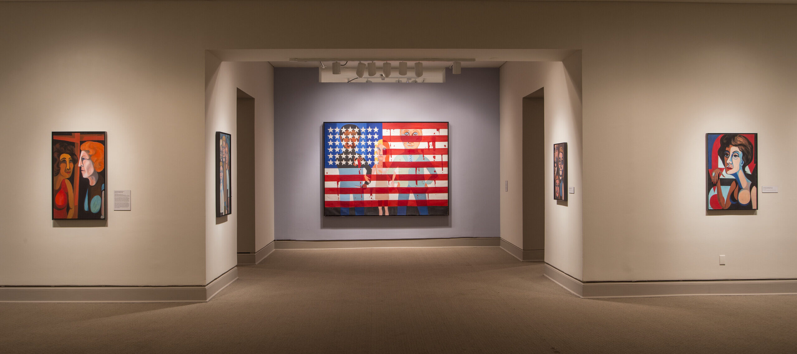 Installation view of a gallery space. A large painting showing an American flag splattered in blood and with people with a light skin tone standing behind the flag is hanging in the middle of the room.