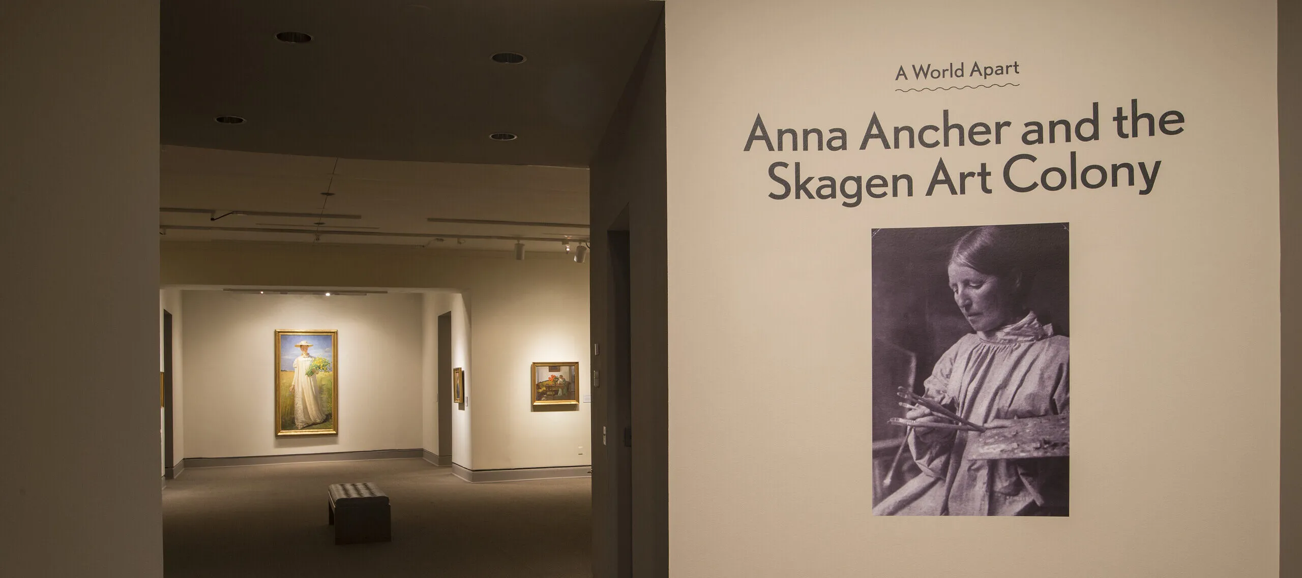 View of a gallery space. On a white wall, it says "A World Apart: Anna Ancher and the Skagen Art Colony."