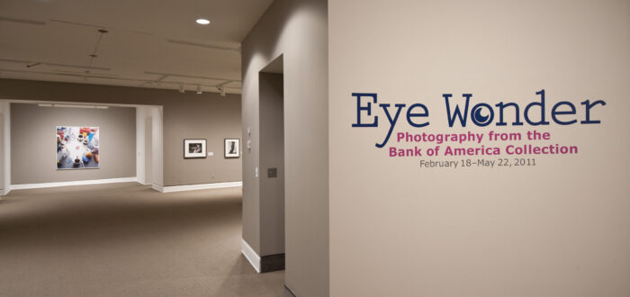 An installation view of a gallery space with white walls and a gray floor. On the wall facing the viewer it says "Eye Wonder: Photography from the Bank of America Collection" in blue and pink letters.