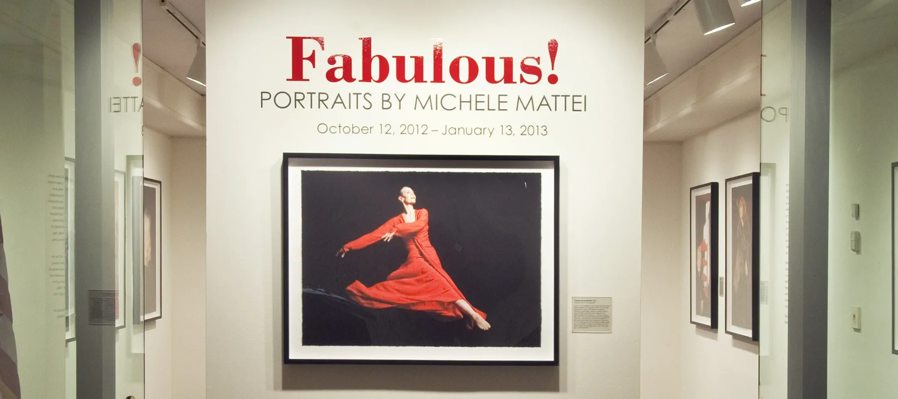 View of a gallery space. On a white wall, a photograph of a woman dancing, wearing a red dress, is hanging below the text "Fabulous! Portraits by Michele Mattei."