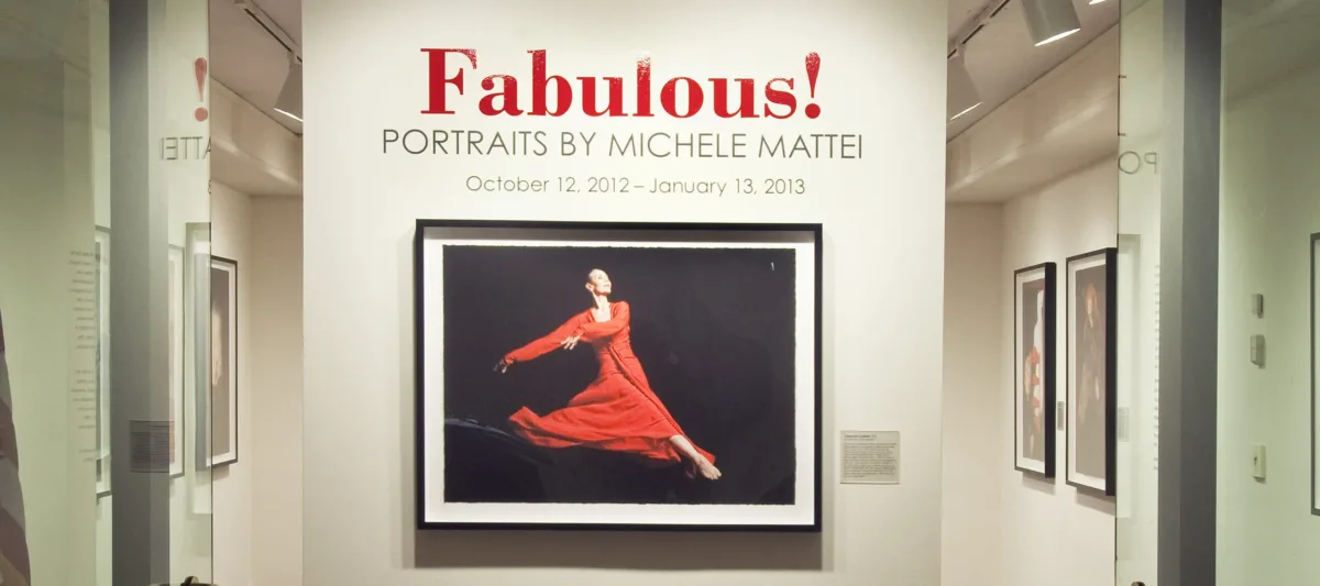 View of a gallery space. On a white wall, a photograph of a woman dancing, wearing a red dress, is hanging below the text "Fabulous! Portraits by Michele Mattei."