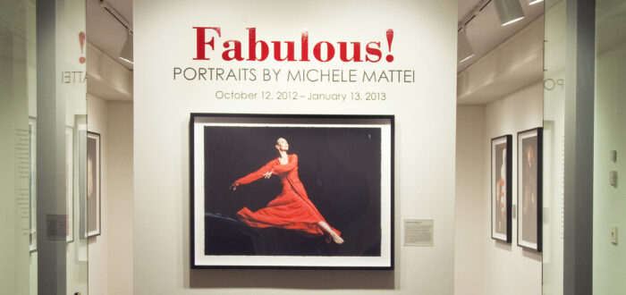 View of a gallery space. On a white wall, a photograph of a woman dancing, wearing a red dress, is hanging below the text "Fabulous! Portraits by Michele Mattei."
