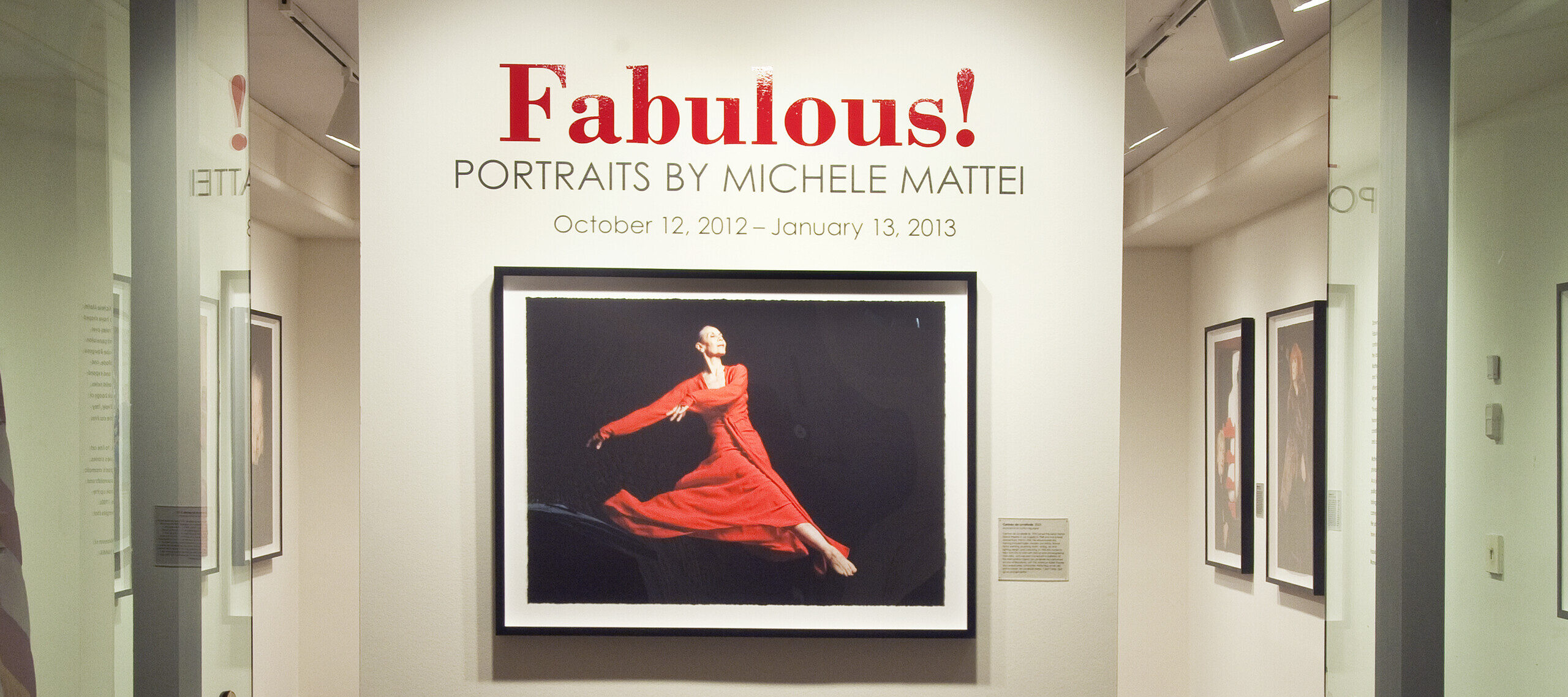 View of a gallery space. On a white wall, a photograph of a woman dancing, wearing a red dress, is hanging below the text 