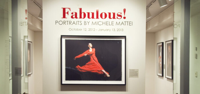 View of a gallery space. On a white wall, a photograph of a woman dancing, wearing a red dress, is hanging below the text "Fabulous! Portraits by Michele Mattei."