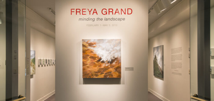 View of an installation. A large painting of a rock formation is hanging on a white wall under the text: "Freya Grand."