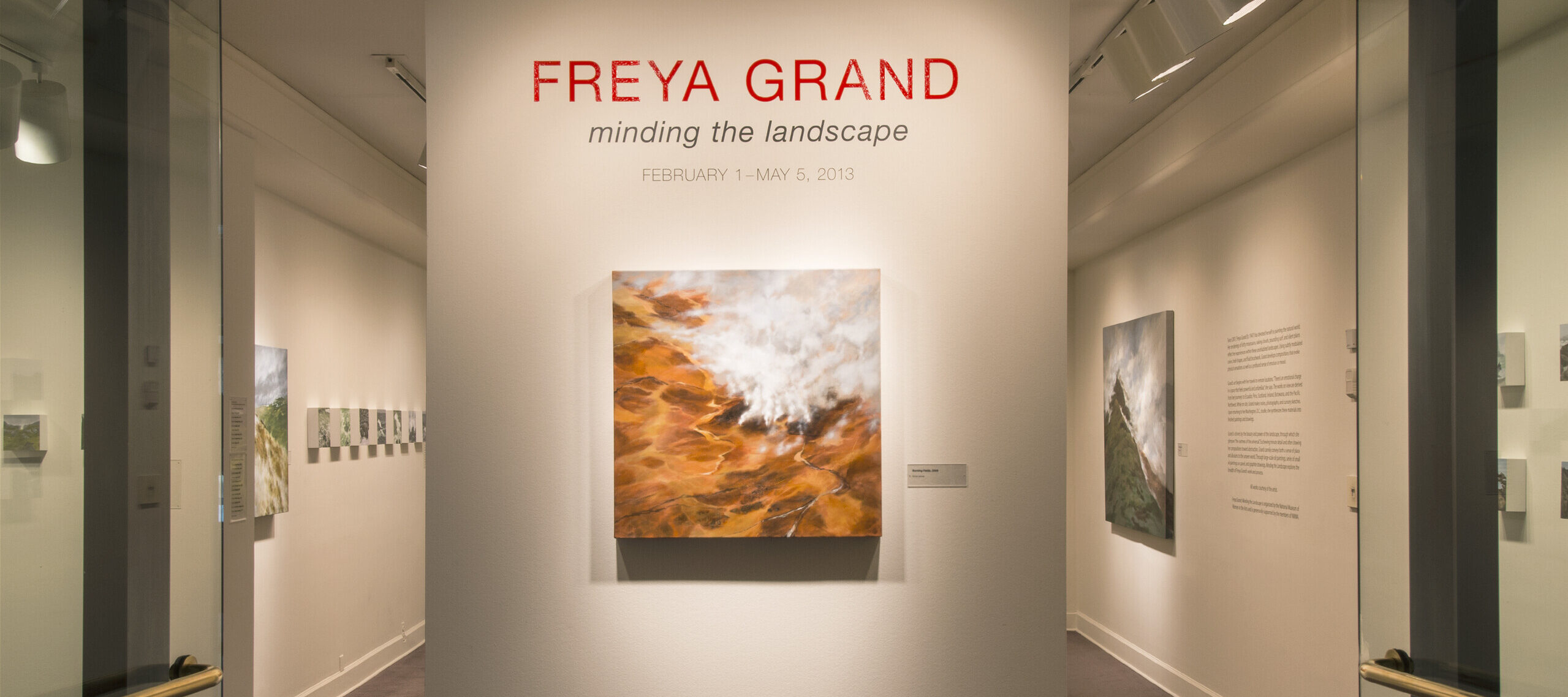 View of an installation. A large painting of a rock formation is hanging on a white wall under the text: 