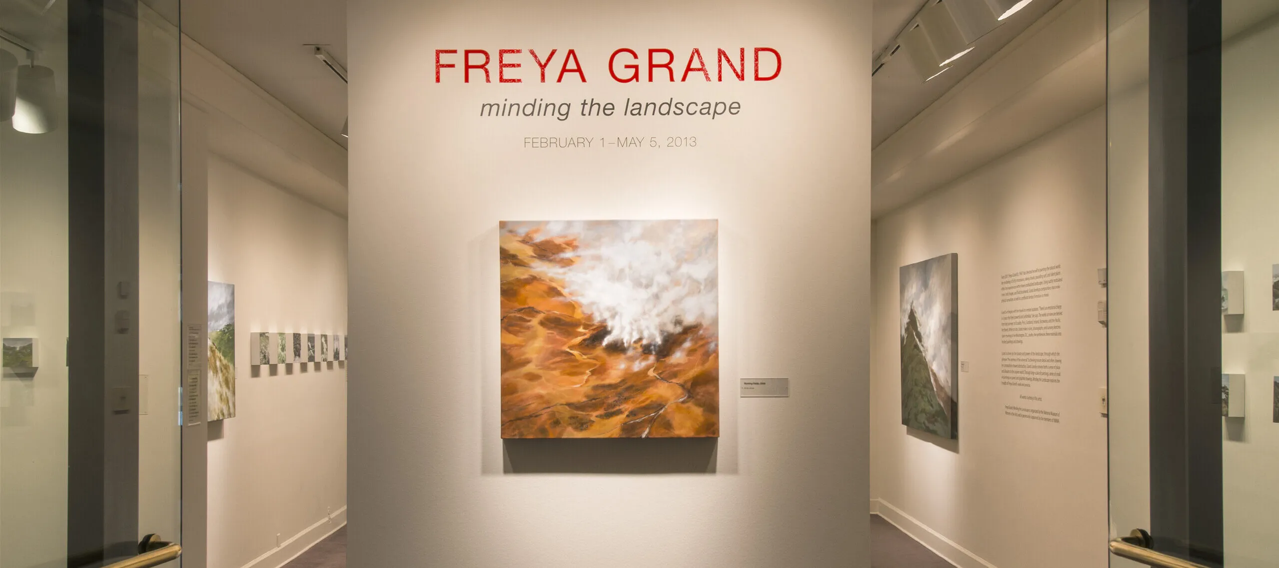 View of an installation. A large painting of a rock formation is hanging on a white wall under the text: "Freya Grand."