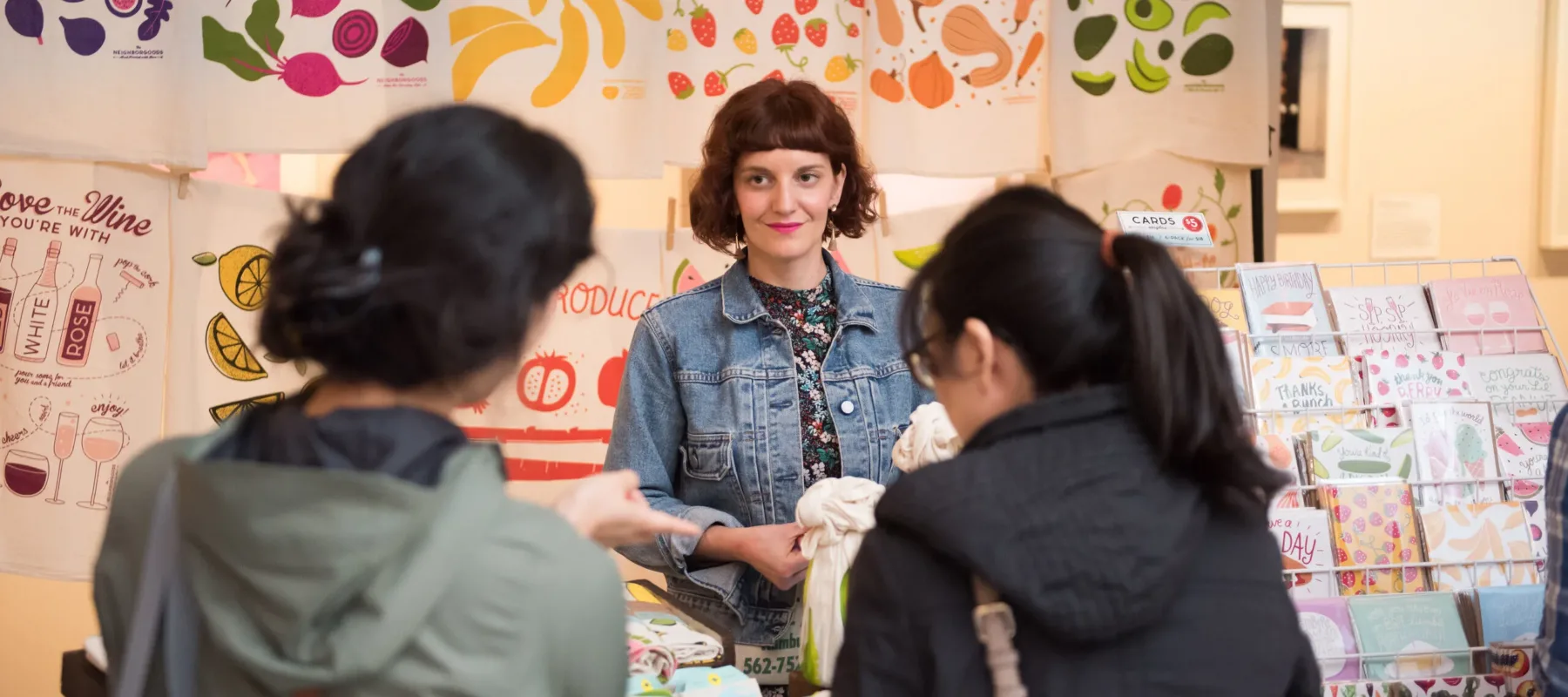 Two people looking at a woman vendor selling colorful dish towels.