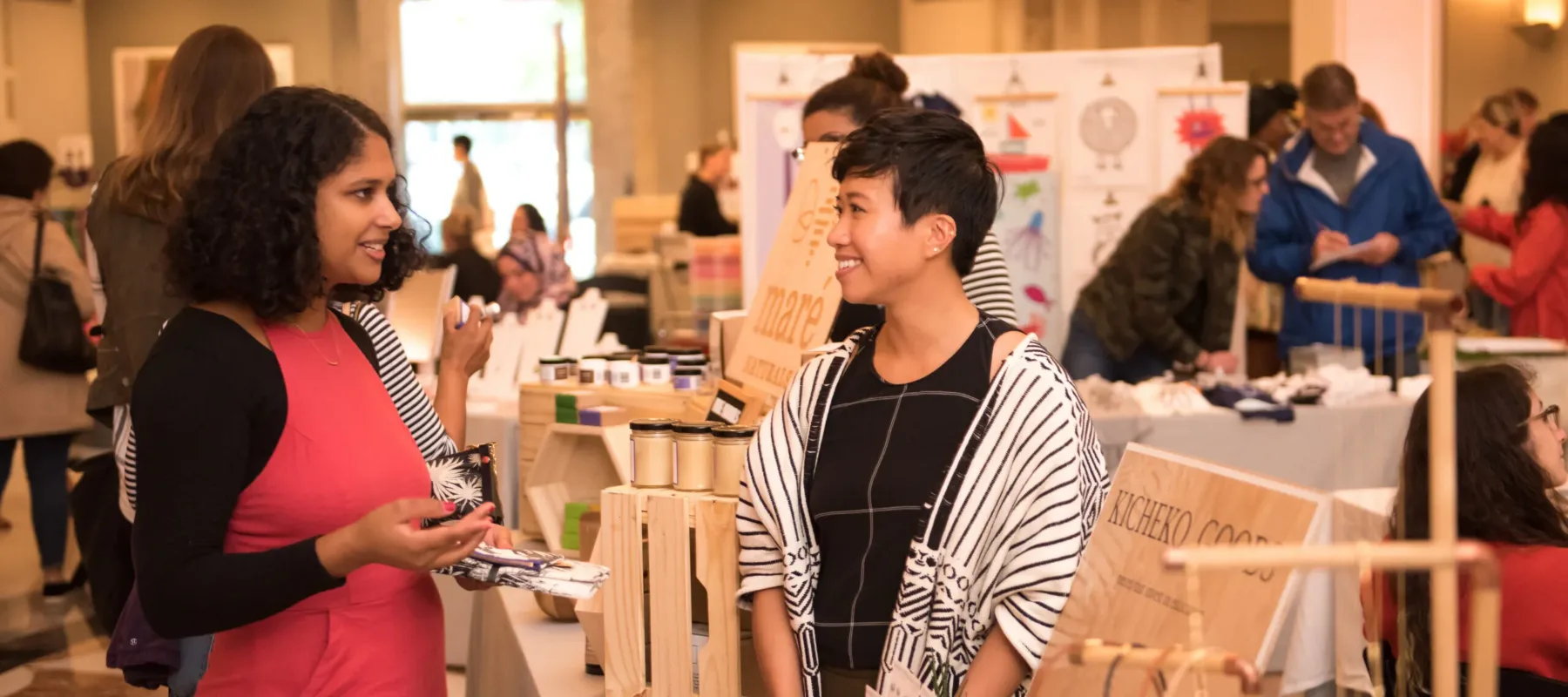 A woman with a medium-dark skin tone wearing a red jumpsuit is talking to a woman with a light skin tone and short, black hair. They are standing in a hall at a fair with people selling handmade goods.