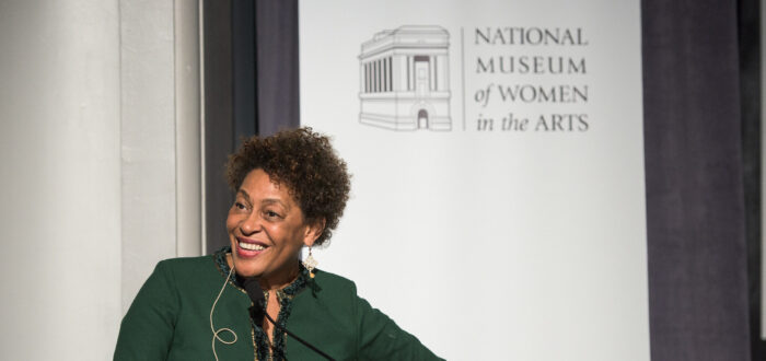 A woman with a medium-dark skin tone and shirt, black hair is giving a lecture before a white banner reading "National Museum of Women in the Arts." She is wearing a forest-green dress and slightly bends her body towards her right.