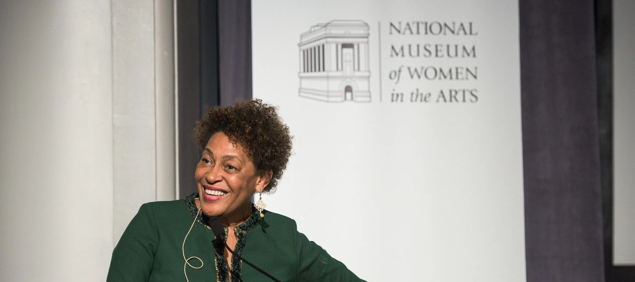 A woman with a medium-dark skin tone and shirt, black hair is giving a lecture before a white banner reading "National Museum of Women in the Arts." She is wearing a forest-green dress and slightly bends her body towards her right.