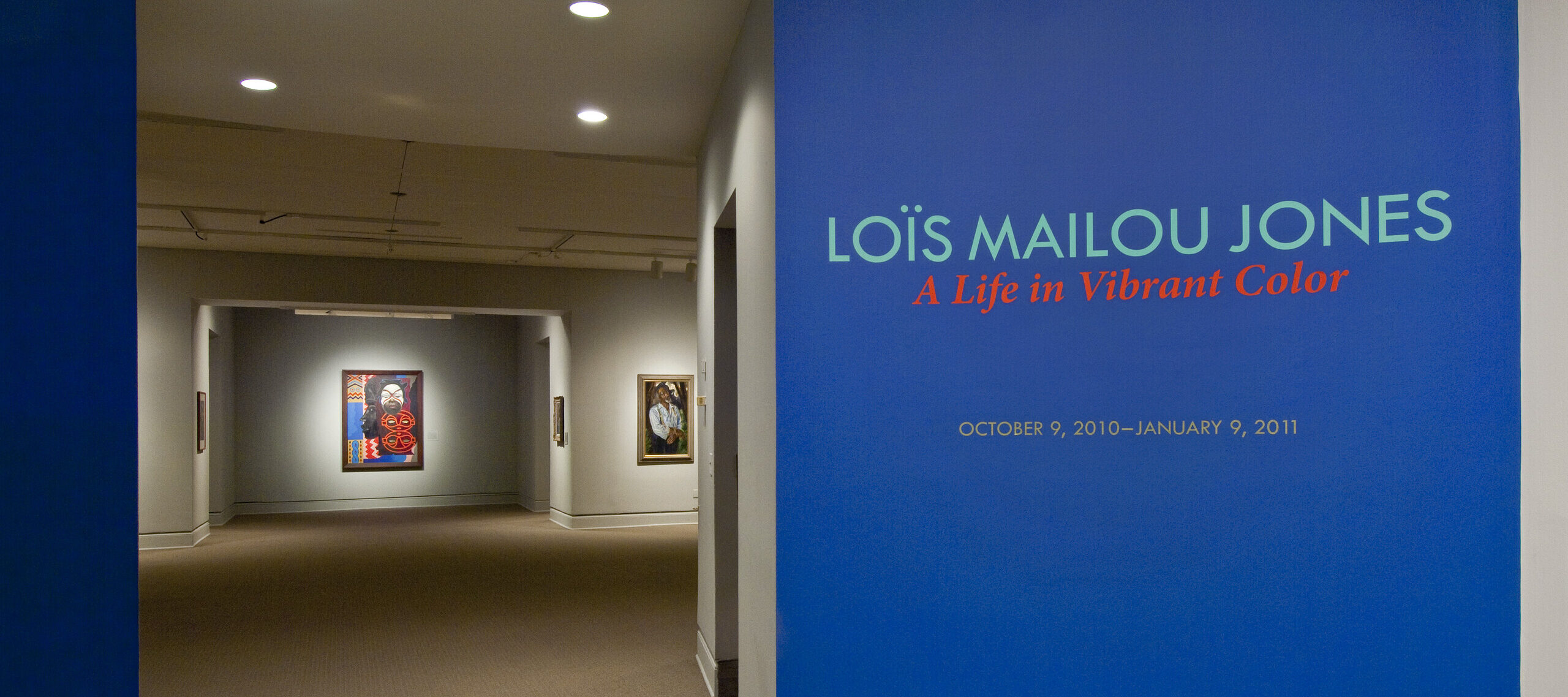 Intervior view of the museum galleries features a vibrant blue wall with the text 'Lois Mailou Jones: A Life in Vibrant Color'. Multiple artworks are displayed in the background.