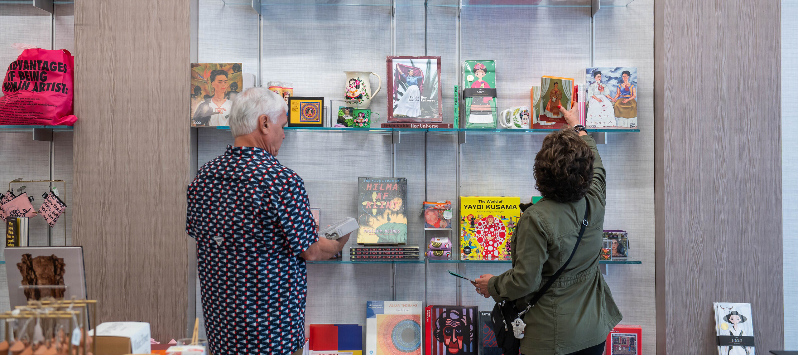 Two visitors are standing in a shop in front wall mounted shelves filled with art books and objects. They have light skin tones and are wearing a patterned shirt and an olive jacket. One of the visitors is reaching for a card depicting a woman in a colorful dress framed by white curtains.