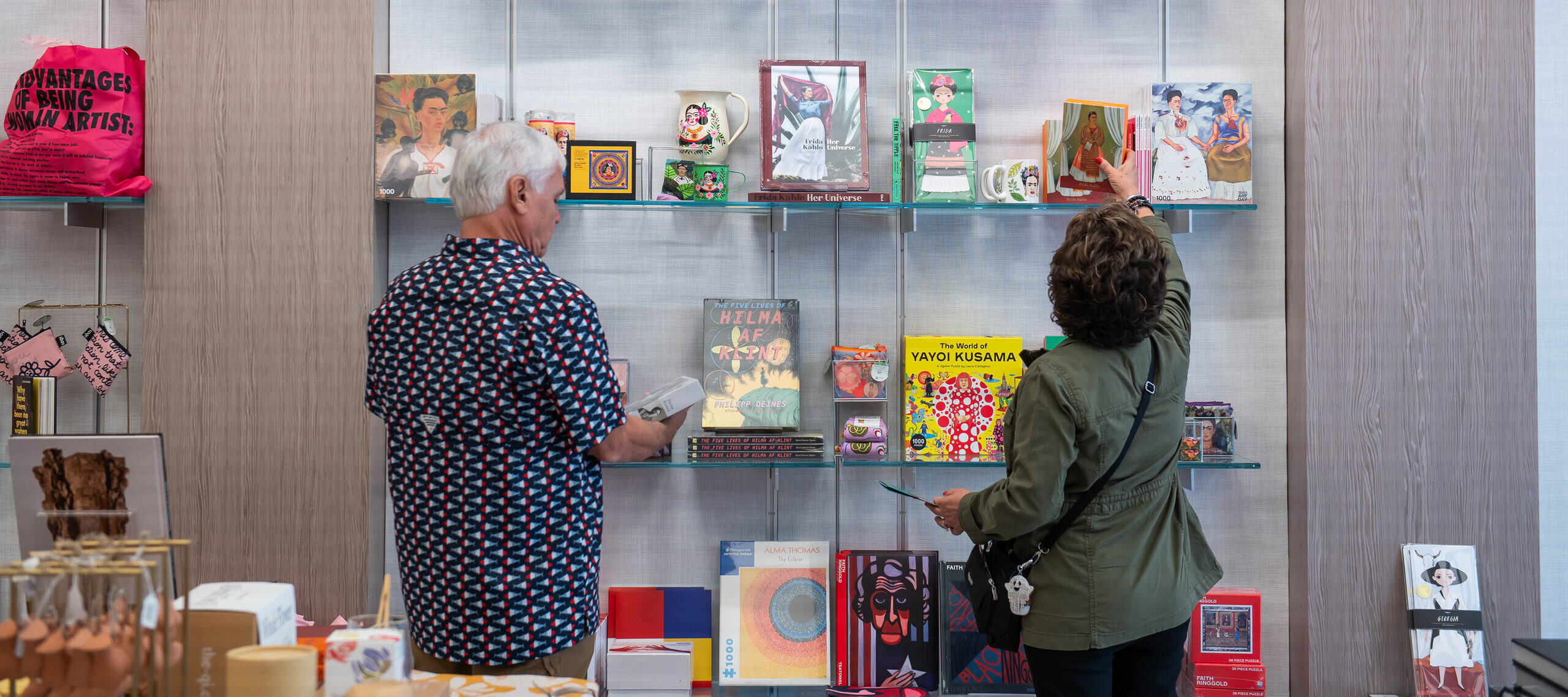Two visitors are standing in a shop in front wall mounted shelves filled with art books and objects. They have light skin tones and are wearing a patterned shirt and an olive jacket. One of the visitors is reaching for a card depicting a woman in a colorful dress framed by white curtains.