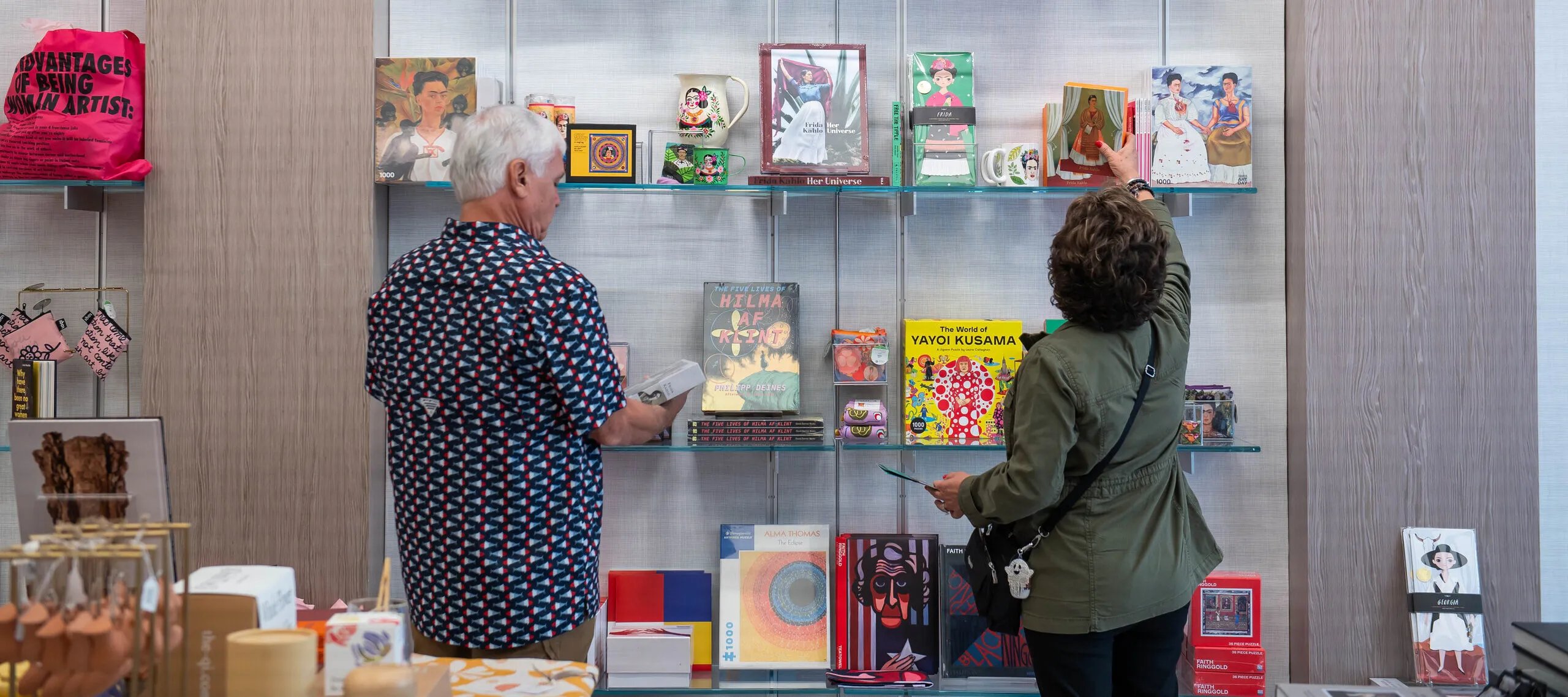 Two visitors are standing in a shop in front wall mounted shelves filled with art books and objects. They have light skin tones and are wearing a patterned shirt and an olive jacket. One of the visitors is reaching for a card depicting a woman in a colorful dress framed by white curtains.